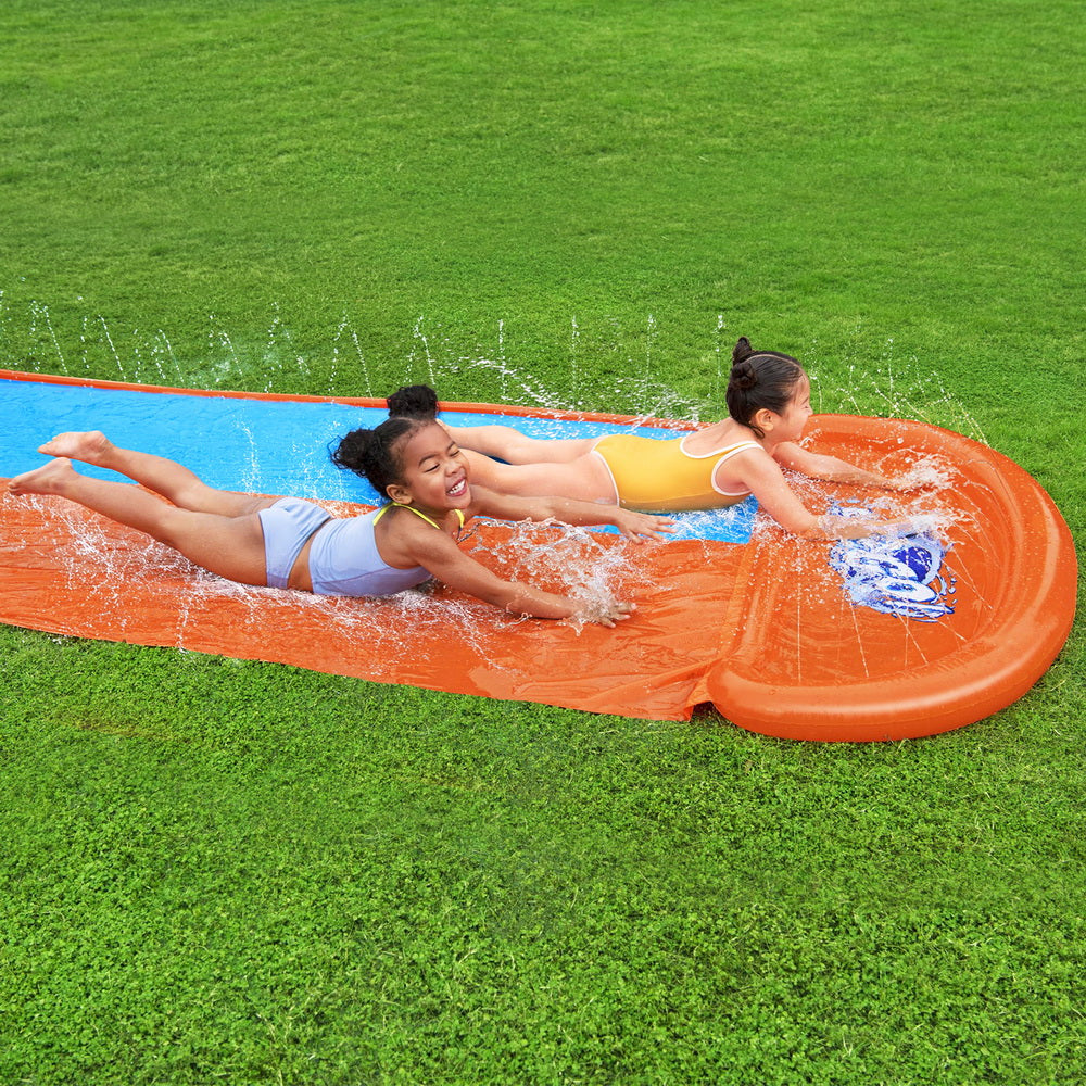 Two kids sliding on orange inflatable splash pool with blue lanes on grass