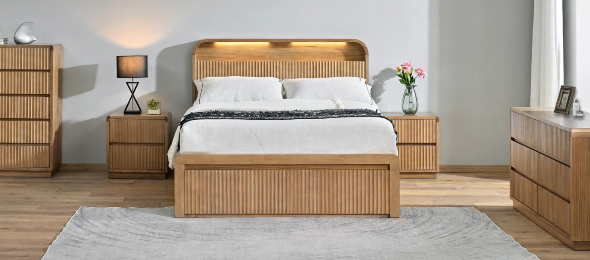 A modern wooden bed frame with a slatted headboard and footboard, styled in a bedroom setting with matching wooden nightstands, a dresser, and a textured rug.