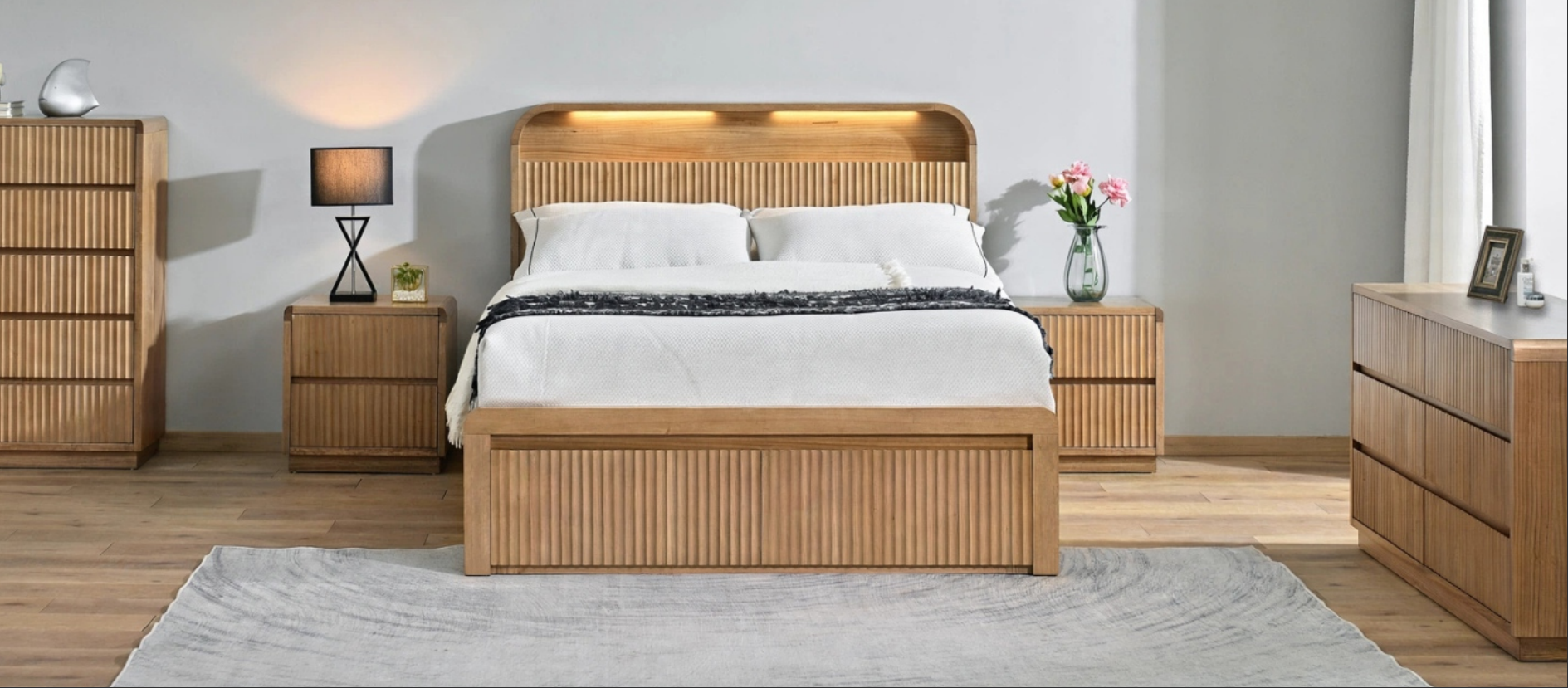 A modern wooden bed frame with a slatted headboard and footboard, styled in a bedroom setting with matching wooden nightstands, a dresser, and a textured rug.