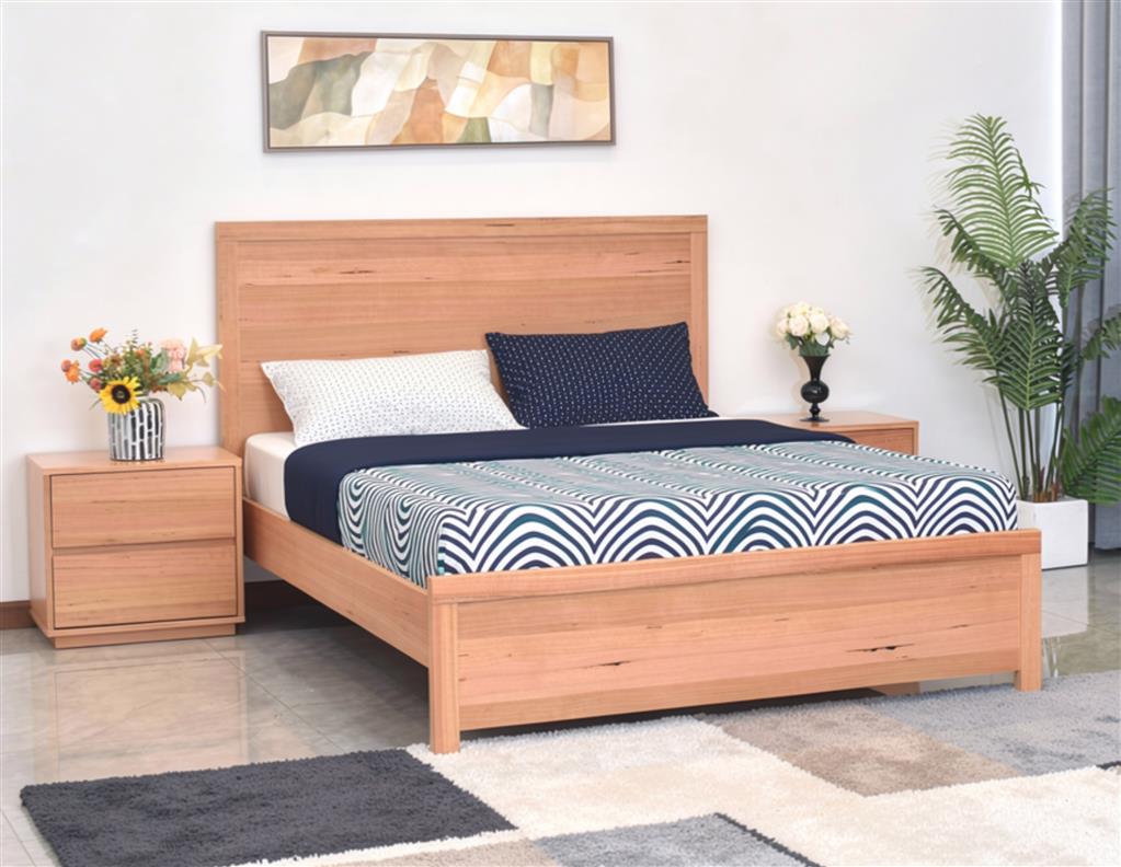 A light oak wood bed frame with a matching headboard and footboard, styled with a blue and white patterned duvet, white and navy pillows, a matching wood nightstand on each side, and a green potted plant in a bright bedroom setting.
