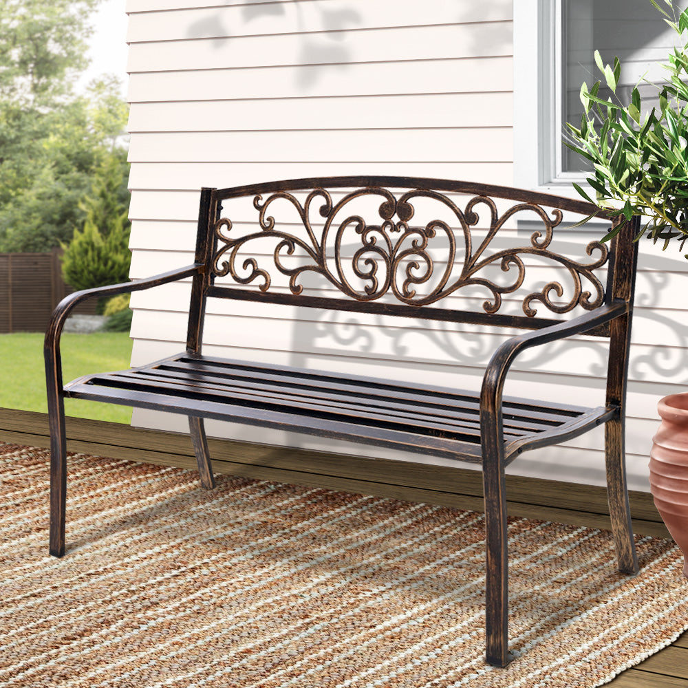 Bronze outdoor garden bench with ornate back panel, curved armrests, and slatted seat