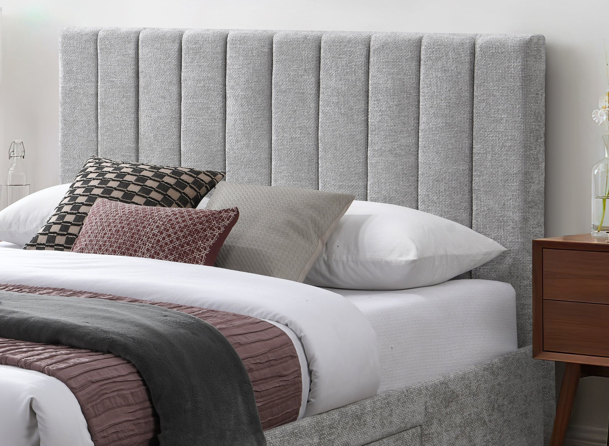 A modern upholstered bed with a tall, vertically channel-tufted headboard in light gray fabric, styled with white bedding, a dark gray throw blanket, and accent pillows on a wooden side table.