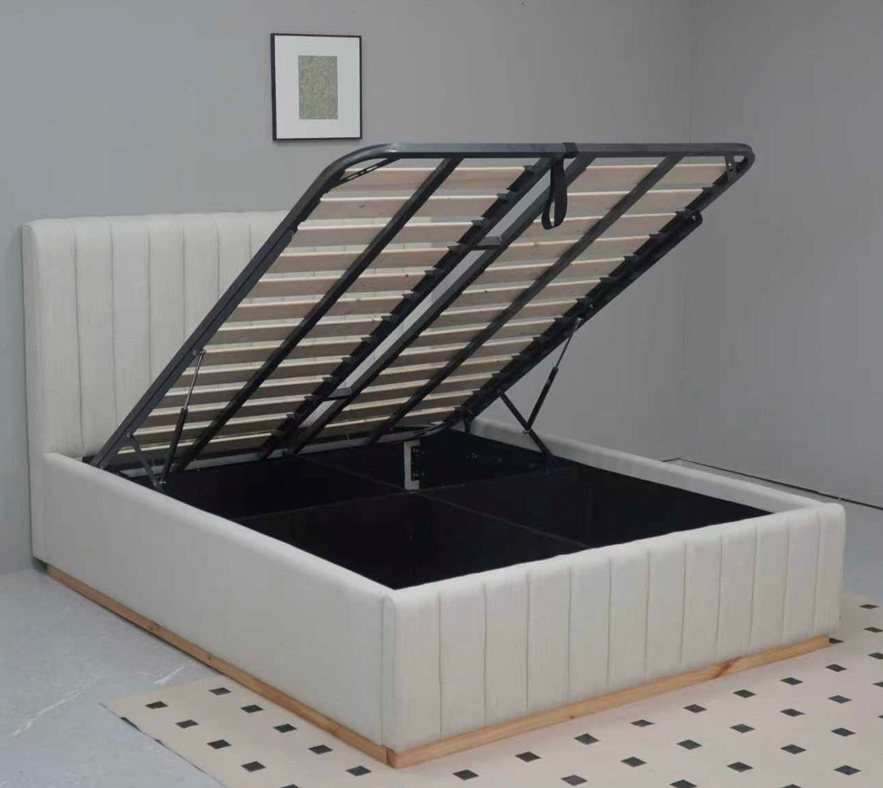 A white upholstered bed frame with the storage compartment open and lid propped up, showing a black slatted support structure. The bed is photographed in a room with light gray walls and a black-and-white checkered floor mat.