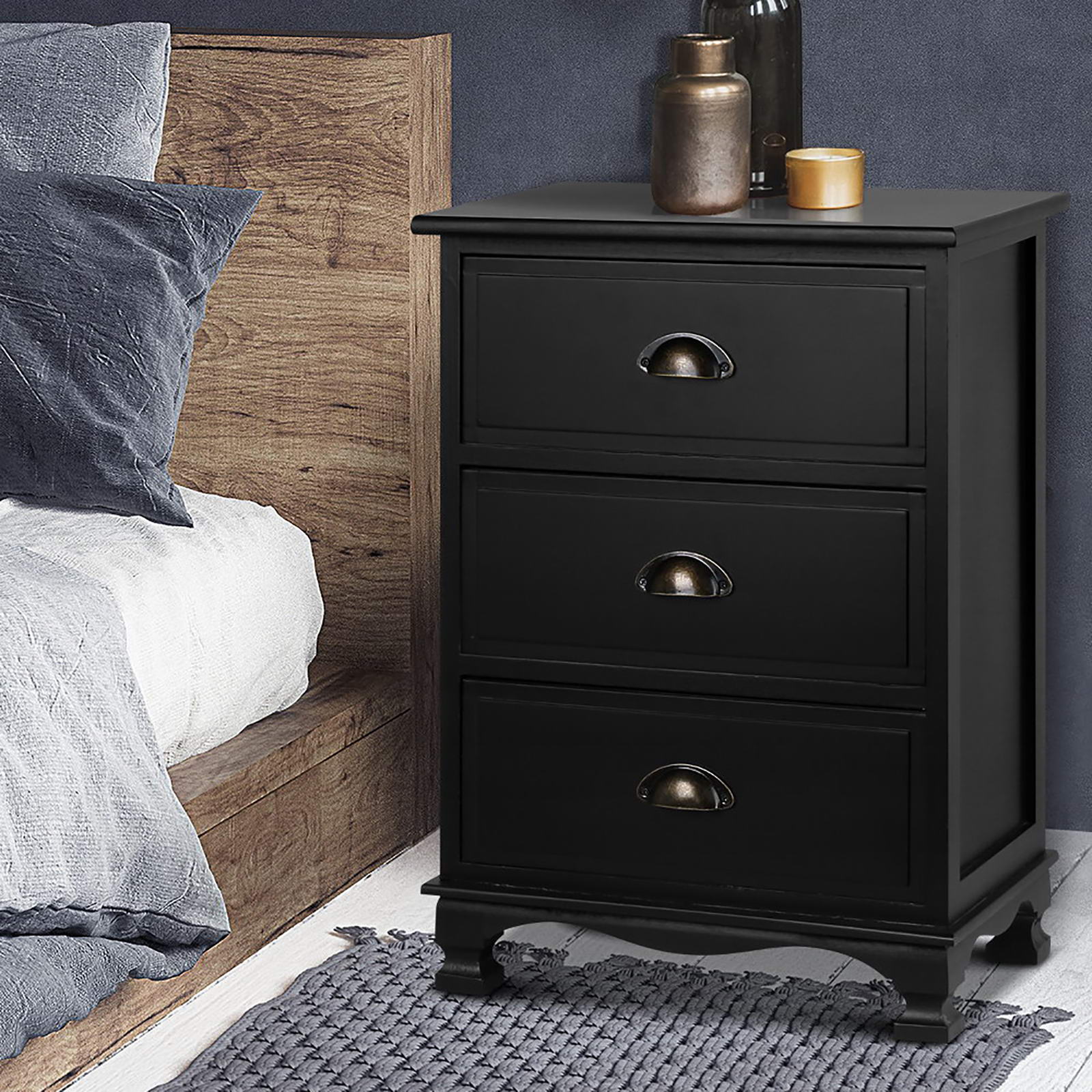Black bedside table with three drawers and arched metal handles, standing beside bed