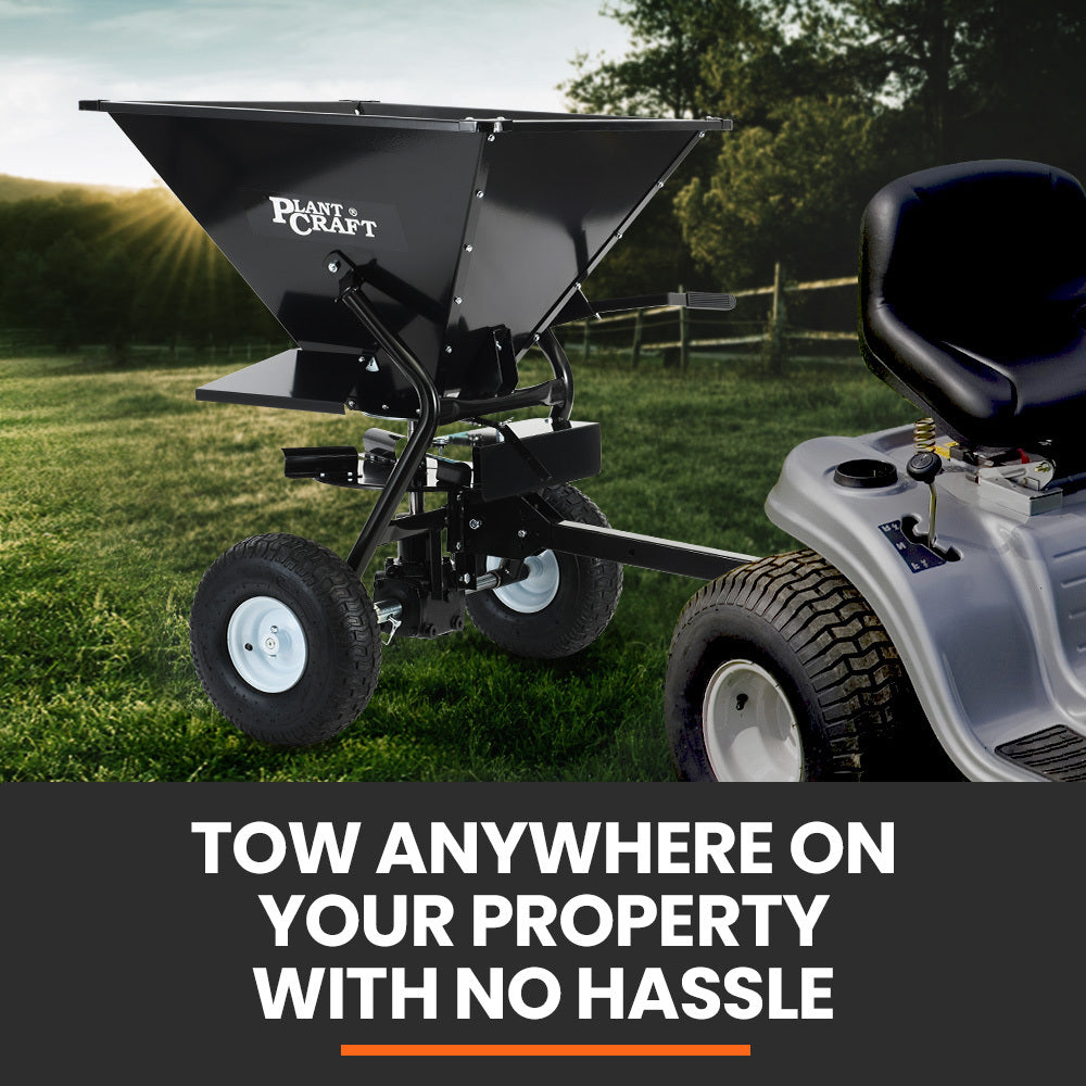 Tow-behind spreader attached to ride-on mower with black hopper and wheels