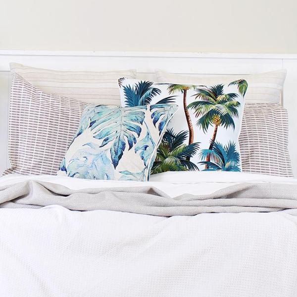 Two white cushion covers with teal palm tree prints on a bed with gray bedding