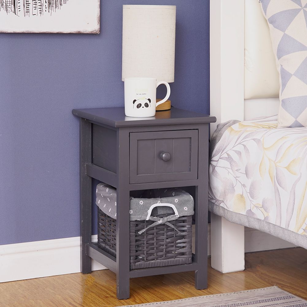 Grey bedside table with drawer and wicker basket, topped with lamp and cup.