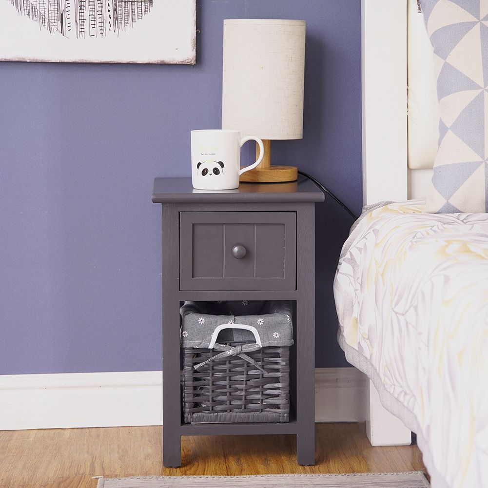 Grey bedside table with drawer and wicker basket, with mug and lamp on top.