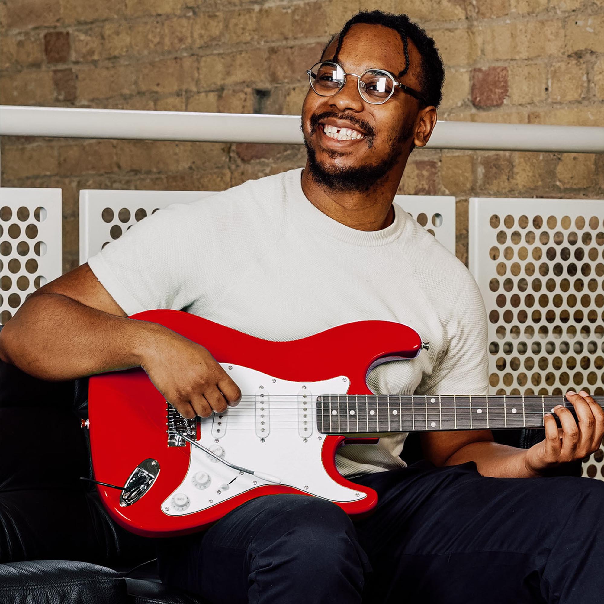 Red electric guitar held while seated, with white pickguard and tuning pegs