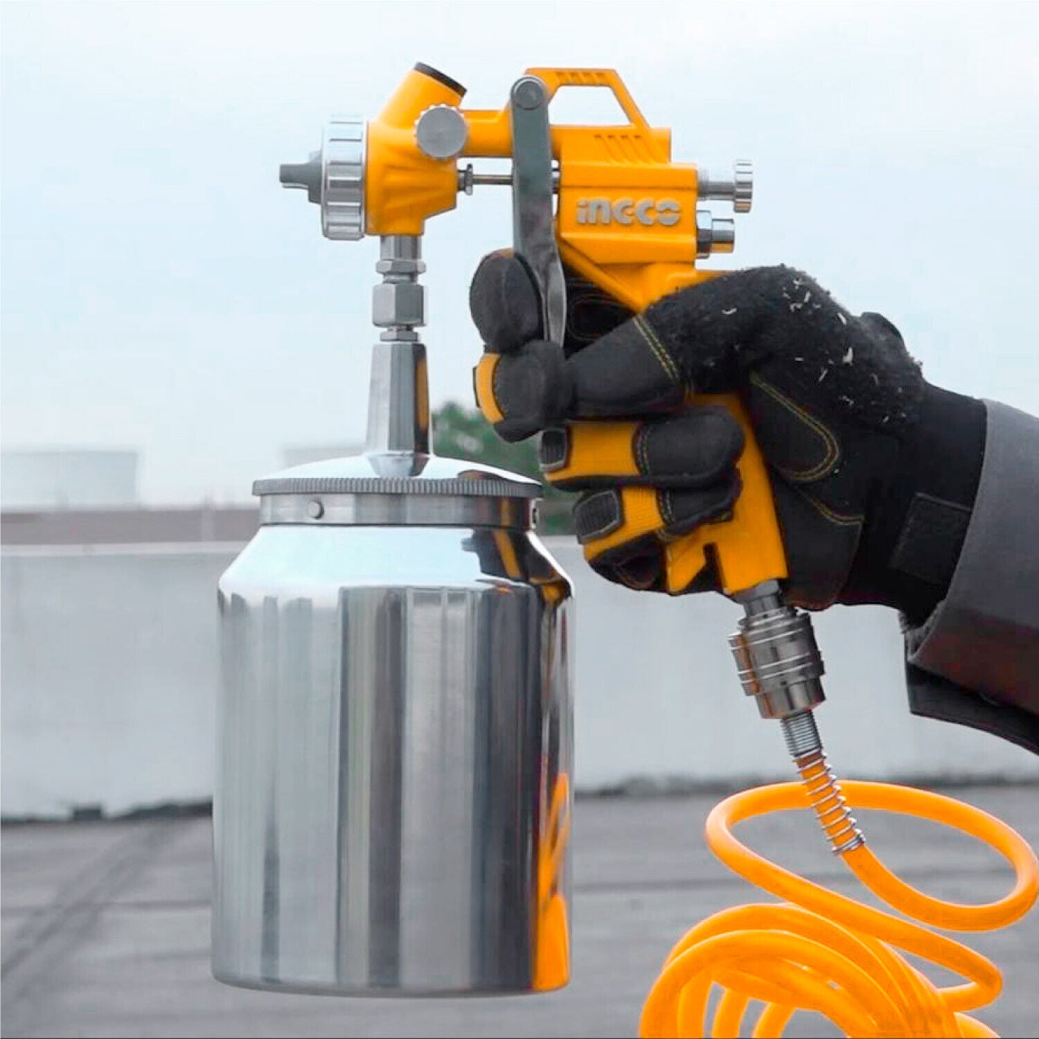 Yellow Ingco paint sprayer with metal cup, held in black gloves