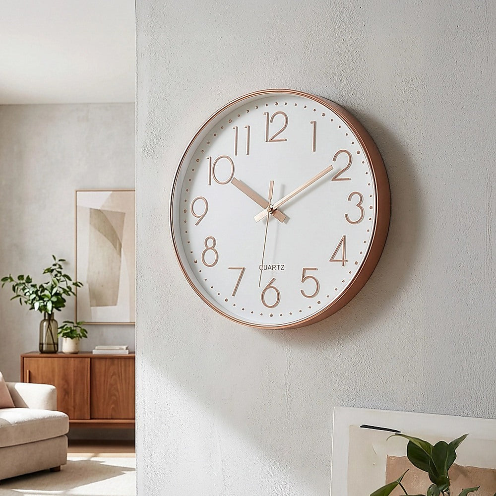 Rose gold wall clock mounted on a wall with white face, roman numerals, and gold hands