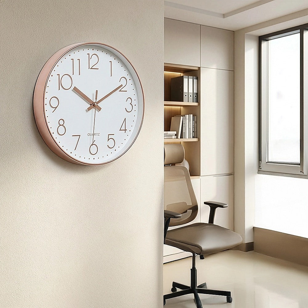 Rose gold wall clock mounted indoors with white face, roman numerals, and gold hands