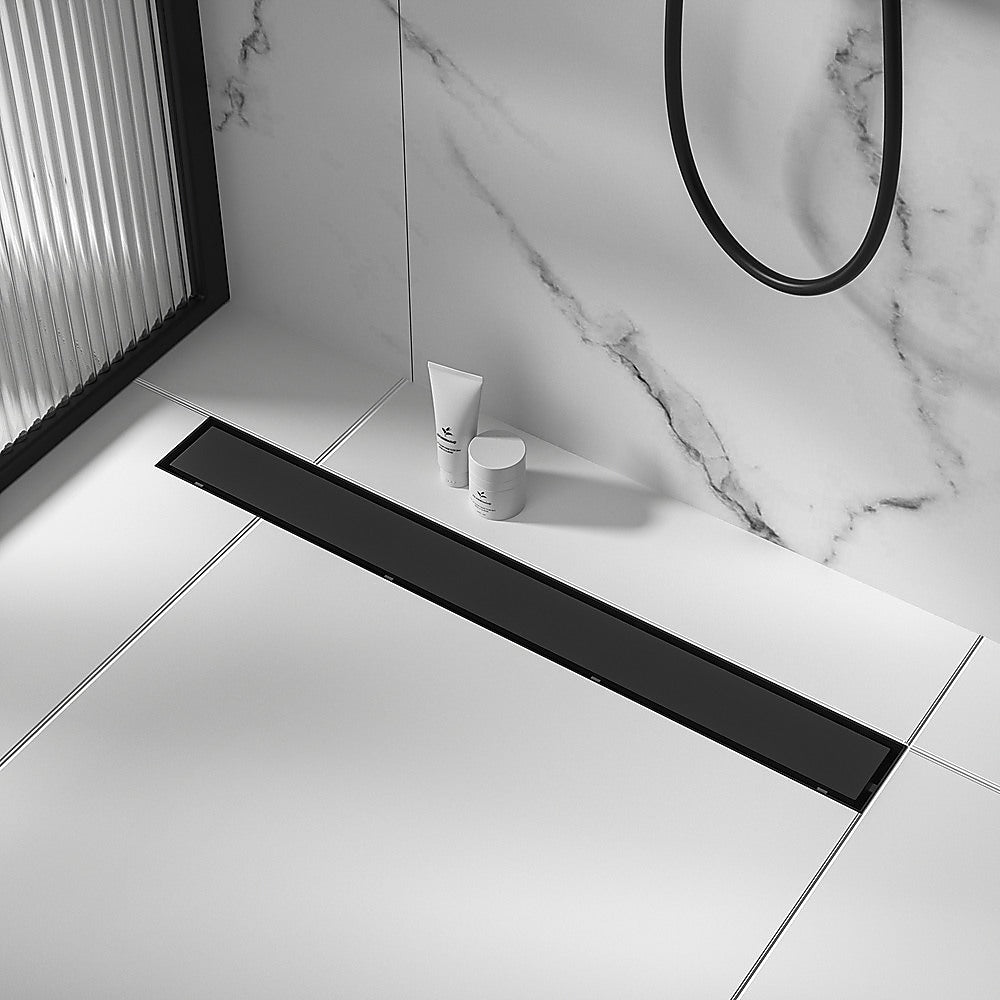 Black linear shower drain grate on tiled floor, long slot and center outlet.