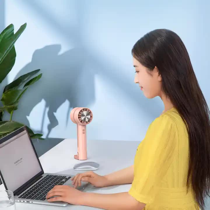 Light pink handheld mini cooling fan on desk stand beside laptop