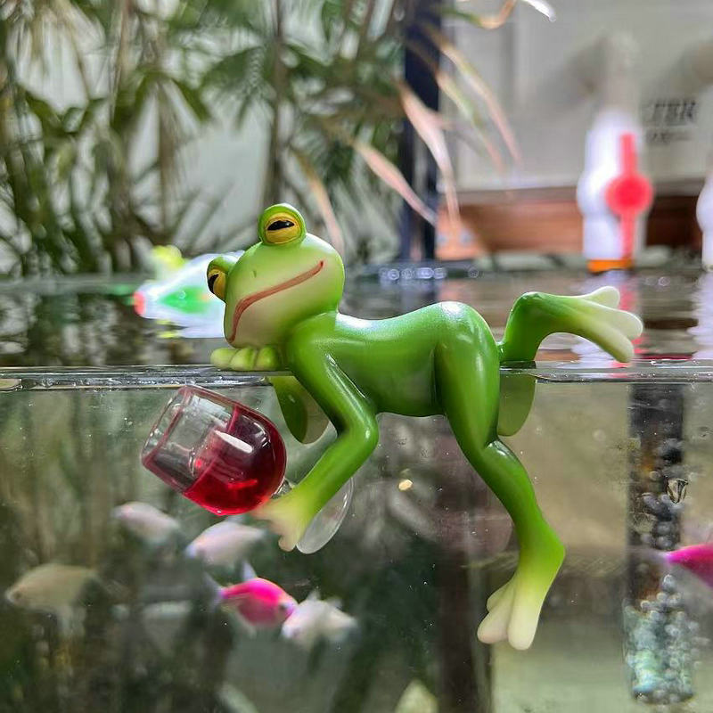 Green frog ornament lying on a glass tank edge with a red hanging mug