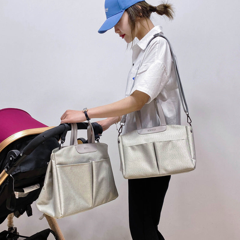 Light gray diaper tote bag worn crossbody, with top handles and front zip pocket panel.