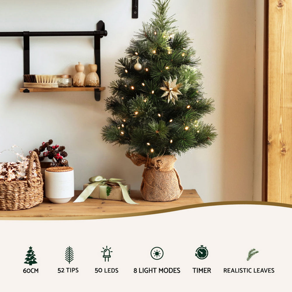 Lit tabletop Christmas tree on a wooden surface with burlap-wrapped base and warm LED bulbs